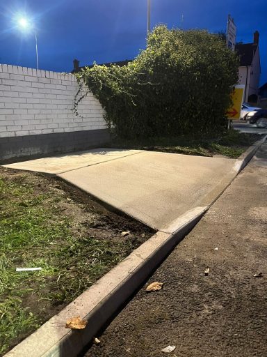 Uneven concrete surface with grass and a bush alongside a dimly lit road at dusk.