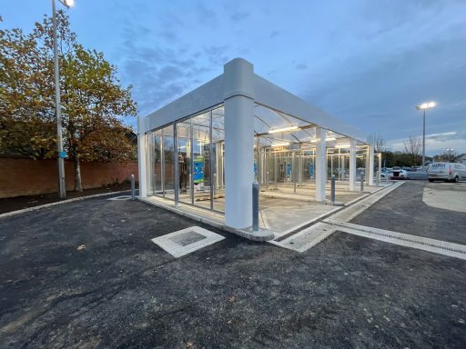 Modern glass building with a clear structure, surrounded by a parking area and trees.
