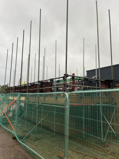 Construction site with vertical steel poles and safety fencing. Overcast sky.